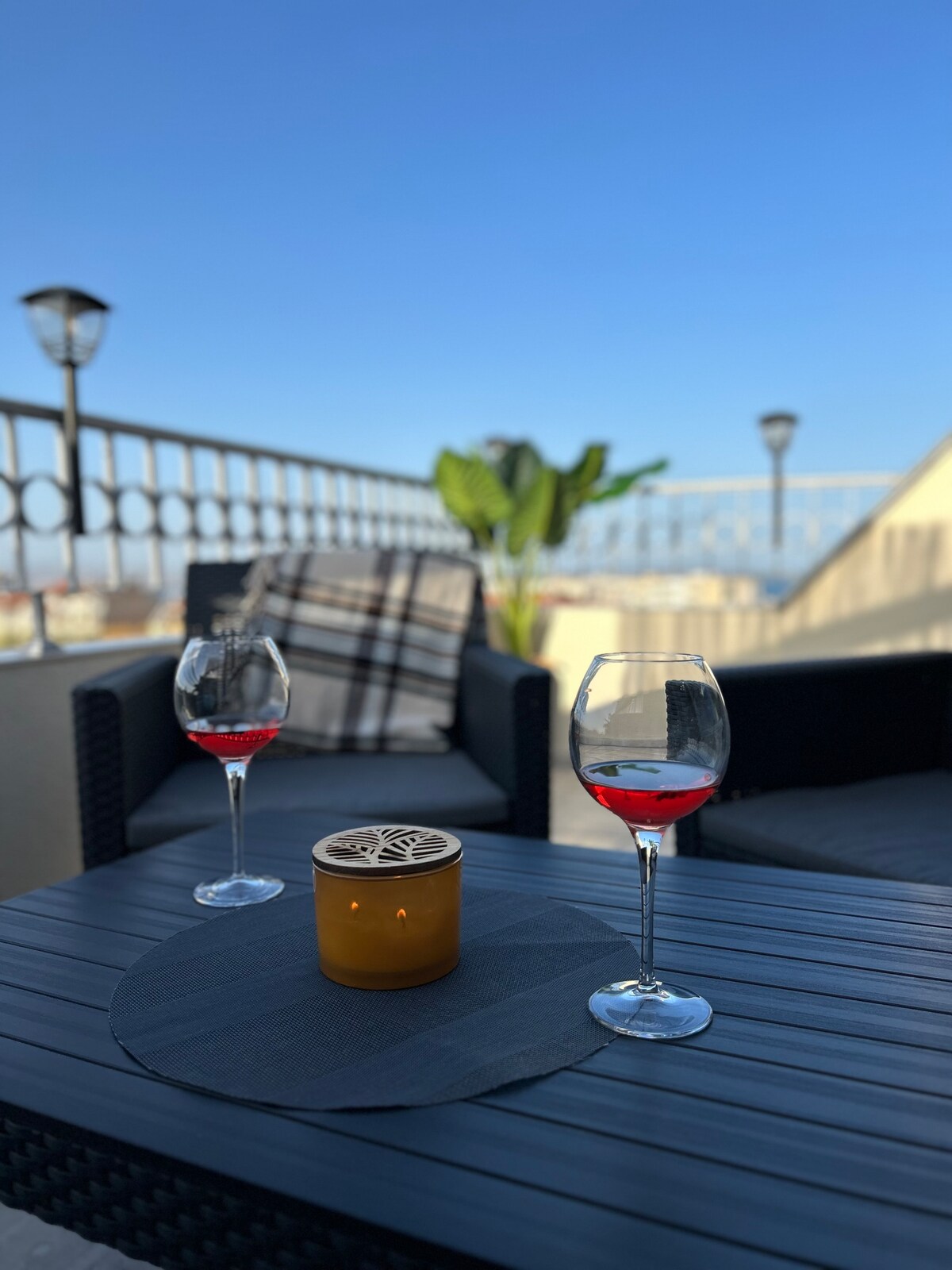 A terrace is shown with two glasses of red beverage placed on a dark round mat atop a table. A lit candle adds a warm glow, while a green plant is visible in the background. Comfortable seating accompanies the serene outdoor setting under a clear blue sky.