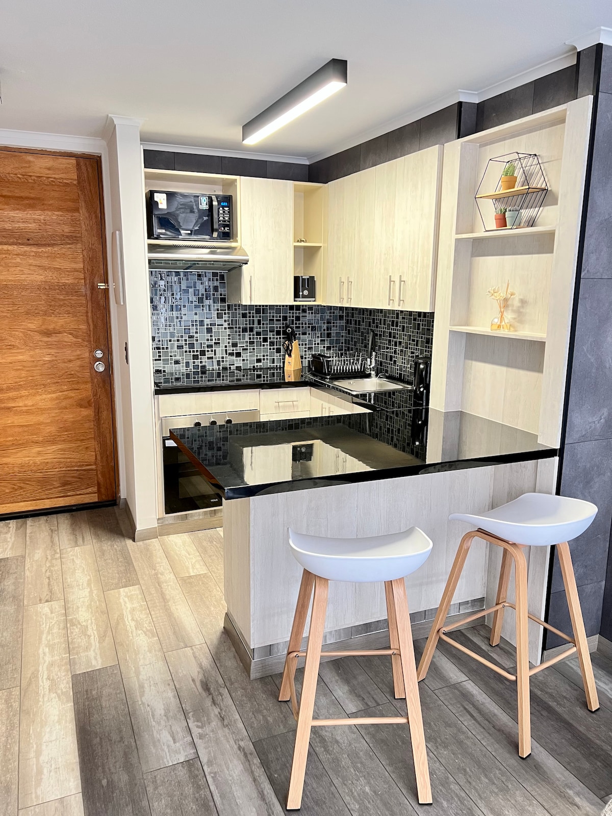 A compact kitchen area features light cabinetry with a dark countertop. An integrated microwave and stove are present, complemented by a mosaic tile backsplash. Two modern bar stools are positioned at the breakfast bar, with light wood flooring adding to the overall aesthetic.