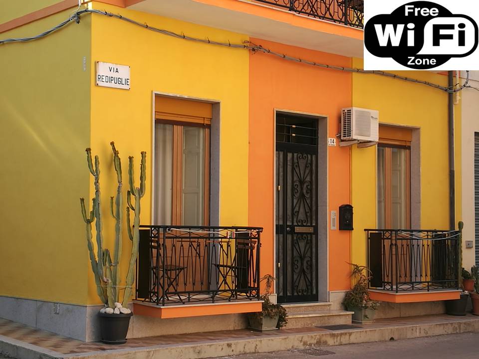The exterior of the apartment features a vibrant yellow facade with decorative wrought-iron balconies. A tall cactus is positioned beside the entrance, which is framed by two windows. A sign indicating 'Via Re D'Ipuglie' is visible above the door. An air conditioning unit is mounted on the wall.