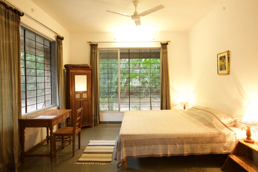 A large room features several windows allowing natural light to filter in, complemented by ceiling fans. A spacious bed is positioned next to a nightstand with a lamp. A wooden desk with a chair is present, and nature views are visible outside.