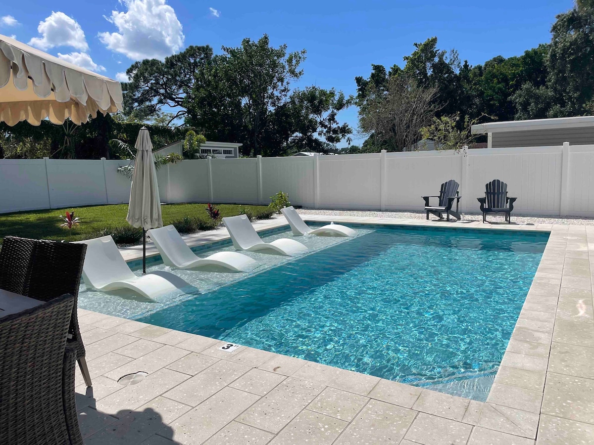 A new pool area features a calm swimming space surrounded by lush greenery. Lounge chairs are positioned partially submerged in the water, providing a relaxing spot. A large umbrella offers shade, and a seating area can be seen nearby under a canopy.