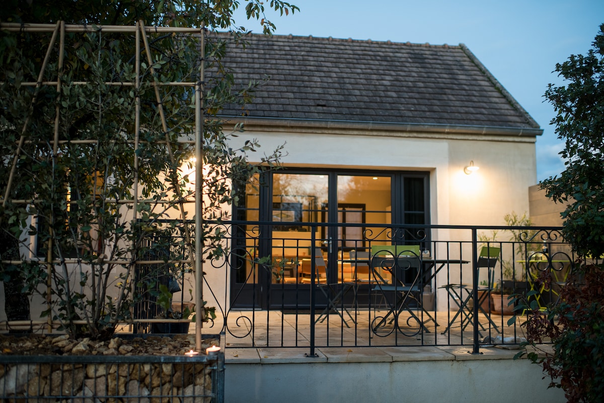 The exterior of the house is shown with a charming terrace featuring black metal railings. Two small chairs and a table are placed outside, illuminated softly by wall-mounted lights. The entrance is framed by greenery, and large glass doors provide a glimpse into the interior.