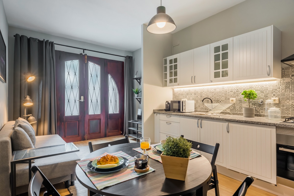 A dining area features a round table set with breakfast items and drinks, complemented by a kitchen with modern appliances and patterned backsplash. A sofa is positioned nearby, and large windows with dark curtains allow natural light to fill the space. The front door adds character with its vintage design.