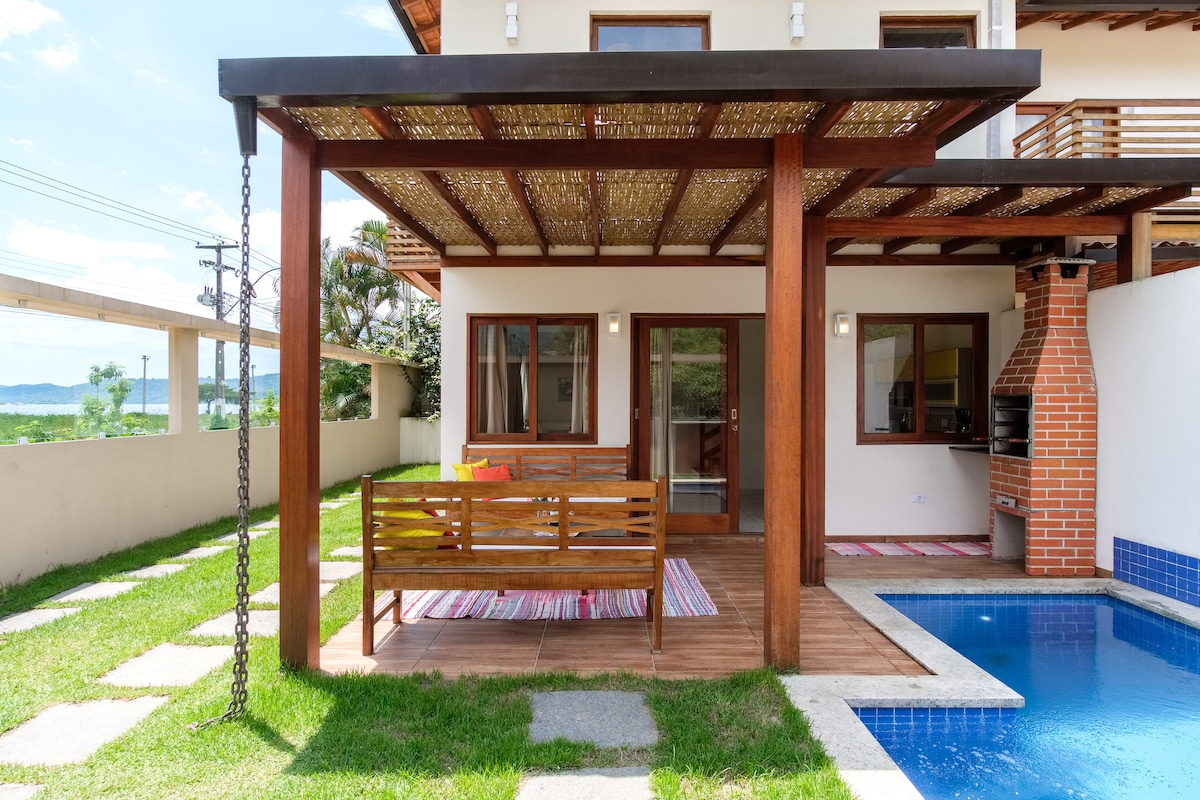 An outdoor patio is shown, featuring a wooden bench and colorful rugs arranged on a wooden deck. A small private pool is visible beside the seating area, surrounded by green grass and a well-maintained garden. A brick barbecue grill is located in the corner.