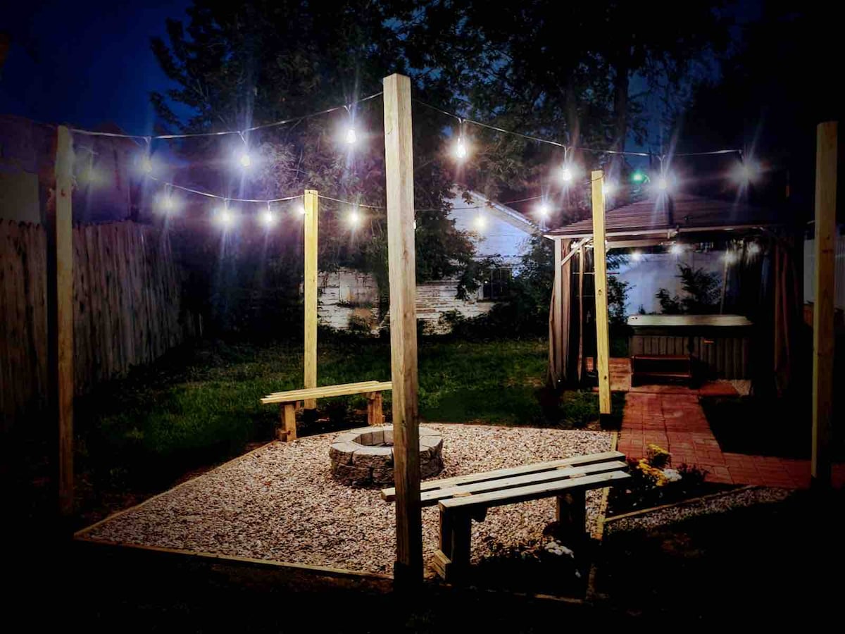 An outdoor space is illuminated by string lights, creating a cozy ambiance in a secluded area. A gravel circle encircles a fire pit, surrounded by handmade wooden benches for seating. Nearby, a gazebo provides shelter, enhancing the inviting atmosphere for gatherings.