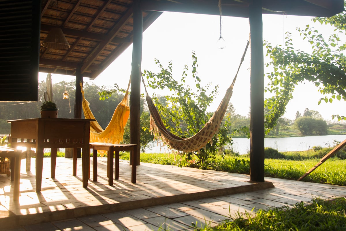 A spacious wooden deck is seen with a wooden table and two hammocks suspended on either side. Soft sunlight filters through the leaves, creating gentle shadows on the tiled floor. The view features a serene body of water in the background, enhancing the tranquil atmosphere.