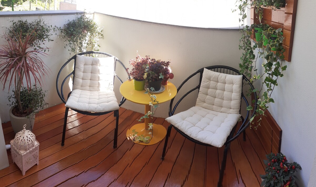 A cozy outdoor seating area is presented with two black chairs featuring plush white cushions, positioned around a small yellow table. Potted plants add greenery, enhancing the tranquil ambiance of the space, which is framed by a wooden deck.