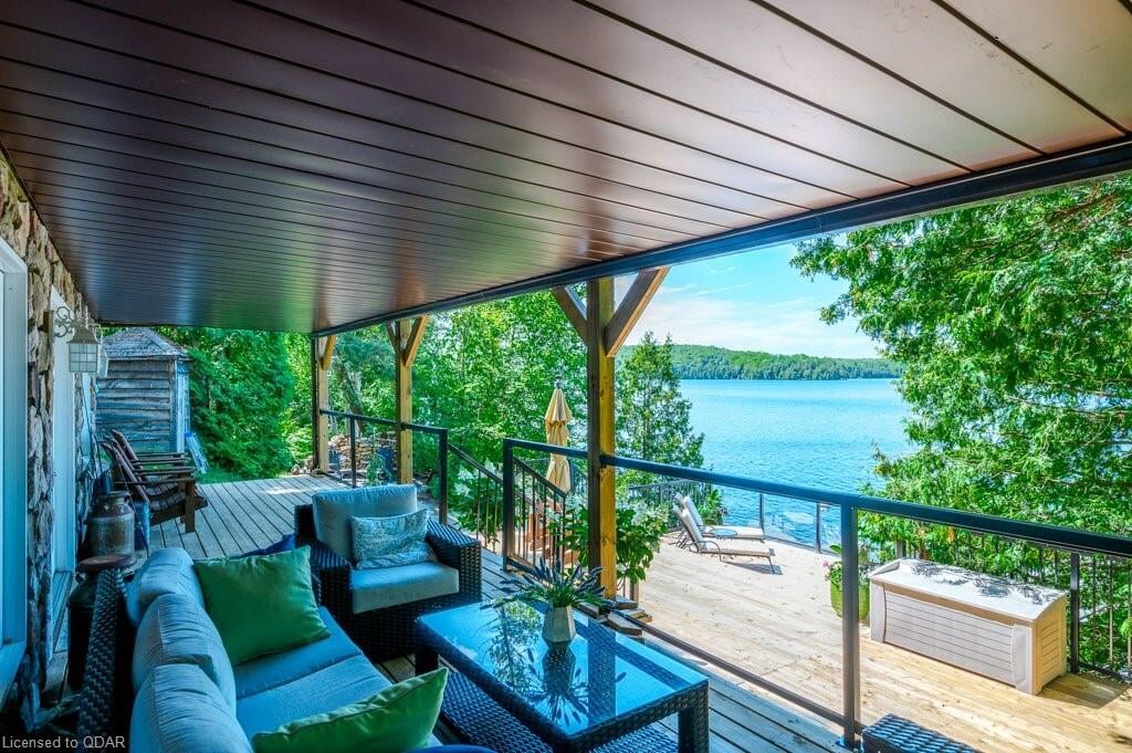 A covered outdoor seating area features a stylish arrangement of furnishings, including a cushioned sectional and a glass-top coffee table. The view beyond showcases the lake and lush greenery, creating a serene backdrop for relaxation.