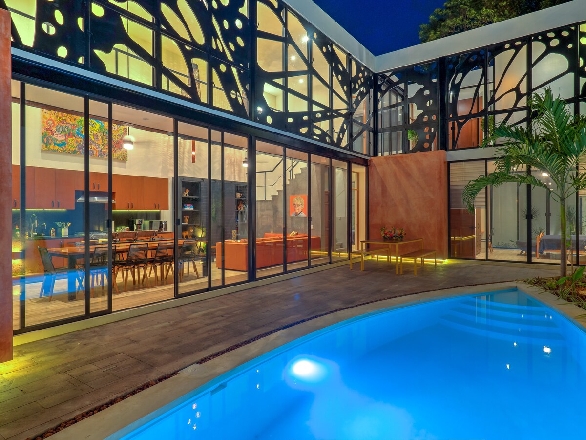 The private patio and pool area is surrounded by striking iron butterfly wings. The outdoor space features inviting turquoise water, with a view of the modern kitchen and dining area through expansive glass doors, creating a seamless connection between indoor and outdoor living.