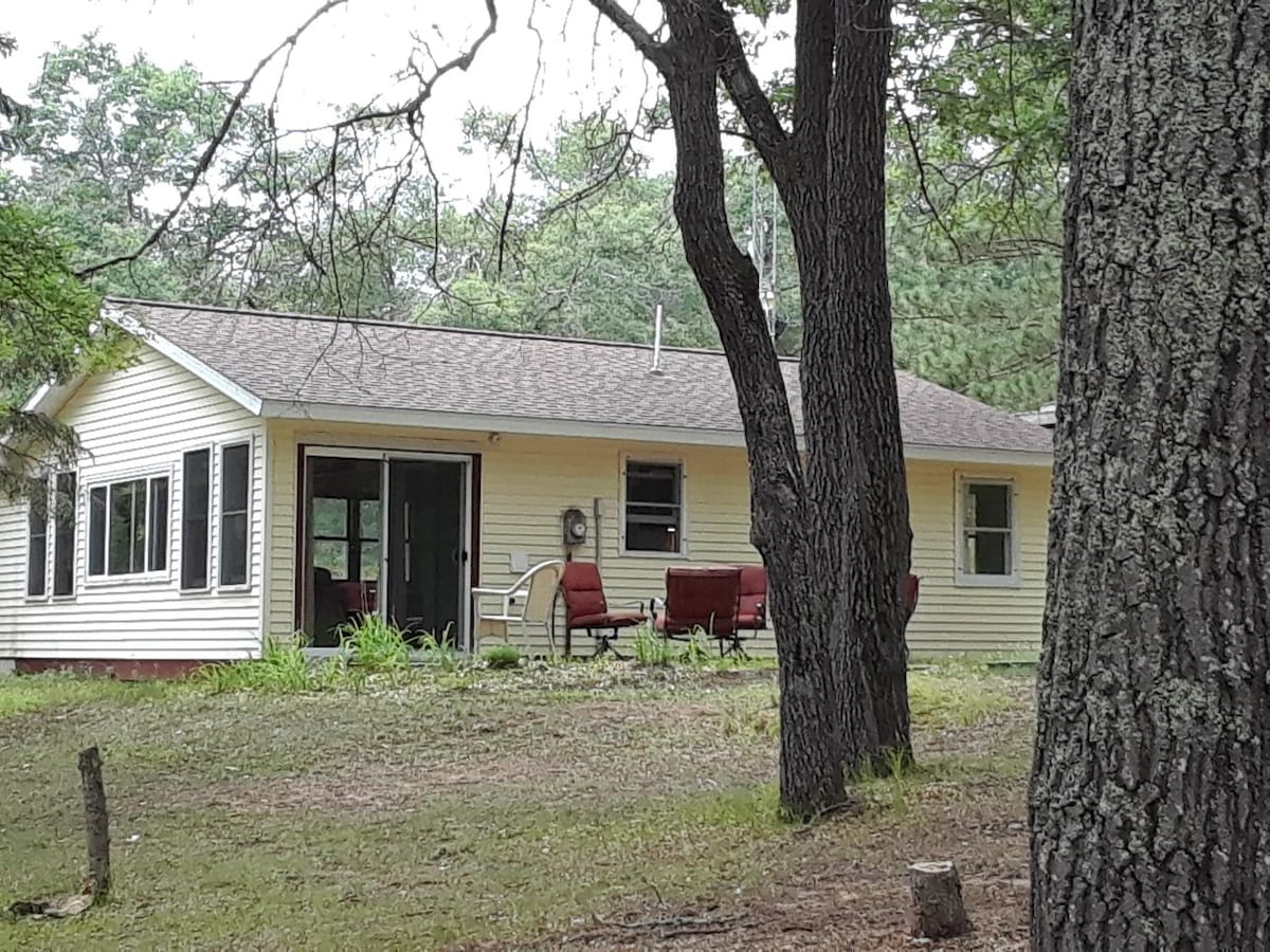 A single-story bunkhouse is surrounded by trees, featuring a spacious patio with red chairs. Large windows allow natural light to fill the interior. The exterior is painted in soft yellow, complementing the natural setting. The peaceful landscape enhances the quiet ambiance.