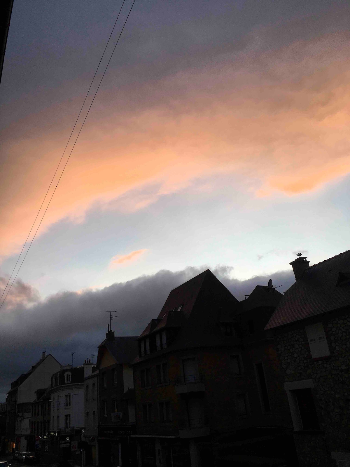 The image captures a scenic view of a neighborhood at dusk, featuring varied rooftops and soft colors in the sky. Warm hues of peach and orange contrast with dark clouds, creating a serene atmosphere as evening approaches. The buildings reflect a typical city structure, showcasing architectural diversity.