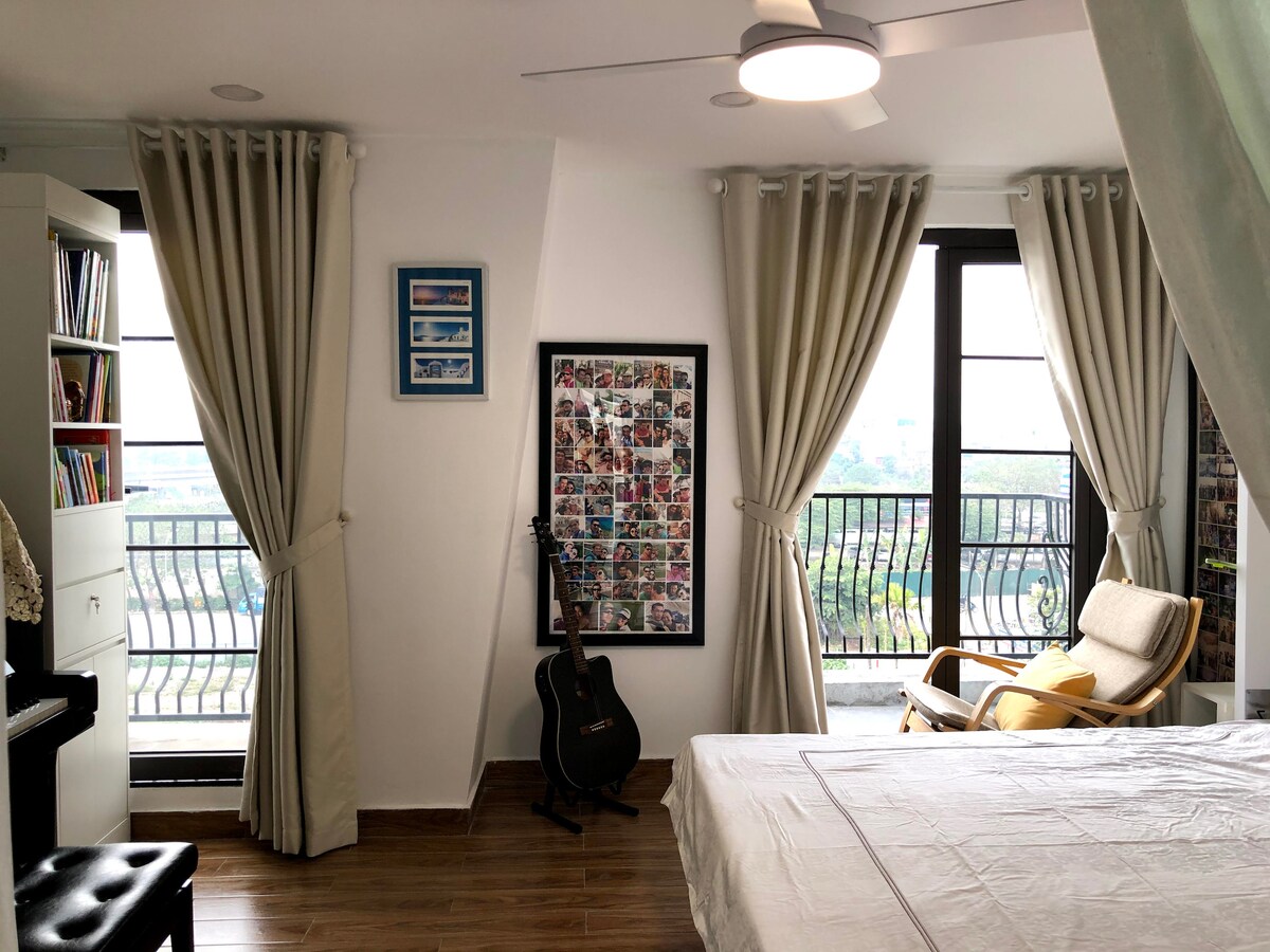 A bright bedroom is shown, featuring large windows with curtains that allow natural light. A spacious bed is positioned near a cozy chair and a bookshelf. A guitar is visible beside the bed, and a piano is located in the corner, adding musical elements to the space.