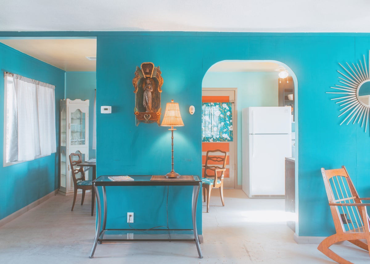 A bright room features vivid blue walls, complemented by warm wooden accents. A small table with a lamp stands near a vintage mirror. Natural light streams in through the window, highlighting the open layout that connects to a kitchen area visible in the background.