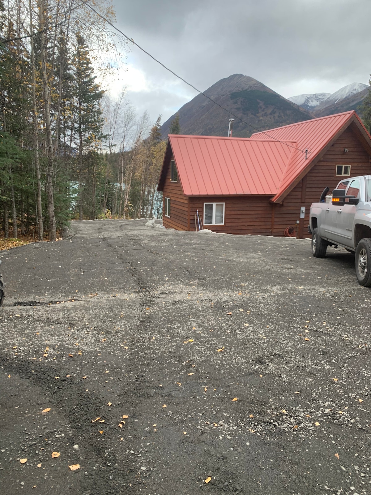 A gravel driveway leads to a rustic cabin with a bright red metal roof. The surrounding area features tall trees and mountains in the background, hinting at the natural beauty of the location. Several vehicles are parked nearby, providing convenience for guests.