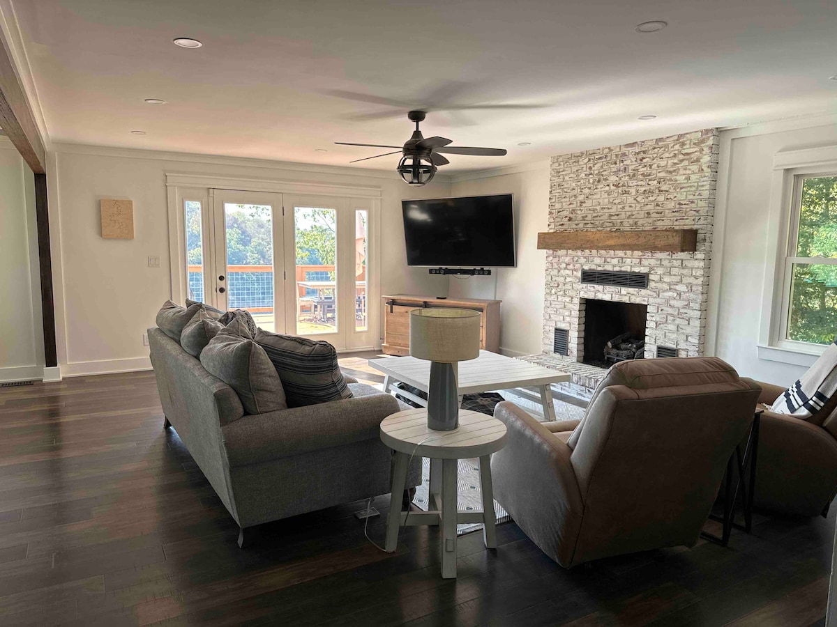 A spacious living area features comfortable seating arrangements, including a gray sofa, two armchairs, and a coffee table. A large flat-screen TV is mounted above a stylish stone fireplace. Natural light fills the space through sliding glass doors that open to a scenic view.