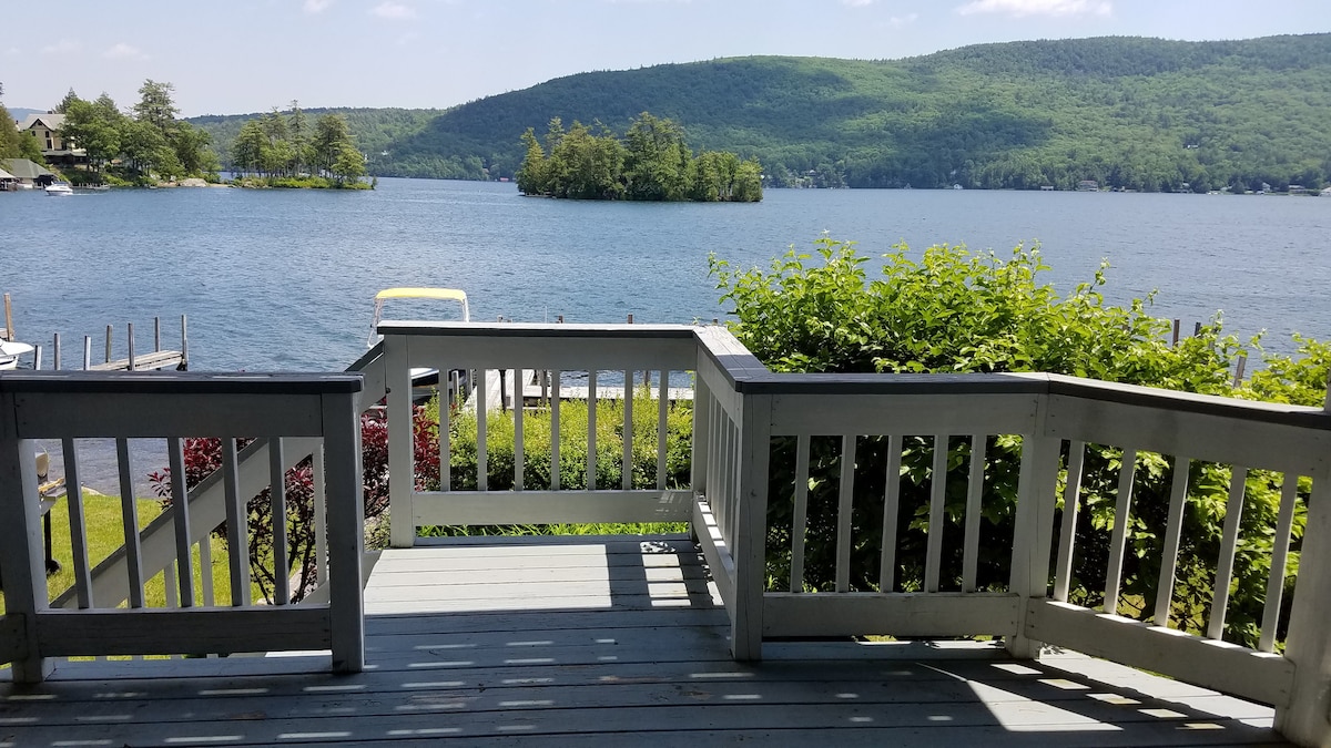 A view from the deck overlooks a serene lake, framed by lush greenery. Gentle slopes and hills in the background enhance the natural setting, while calm waters reflect the surrounding landscape.