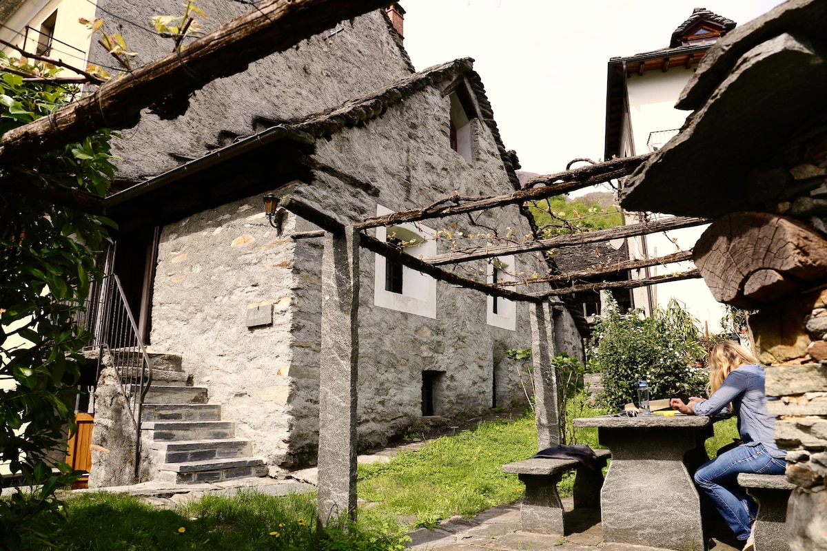 A rustic stone house is surrounded by greenery, with a sturdy wooden pergola overhead. A table made of stone is positioned in the garden, providing a serene spot for meals. The steps leading to the entrance are clearly visible, framed by the natural landscape.
