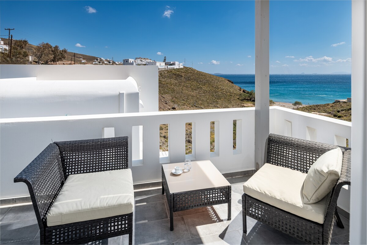 A covered outdoor terrace features two wicker chairs with cushioned seats, positioned around a small table. The view overlooks a serene shoreline with blue waters and distant hills, offering a peaceful place to relax and enjoy the surrounding landscape.