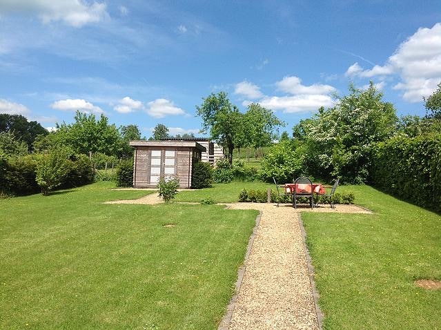 A spacious garden is shown, featuring a well-maintained lawn bordered by trees and shrubs. A gravel path meanders through the greenery, leading to a storage shed. Two chairs are arranged near a small garden with colorful plants, offering a peaceful outdoor space.