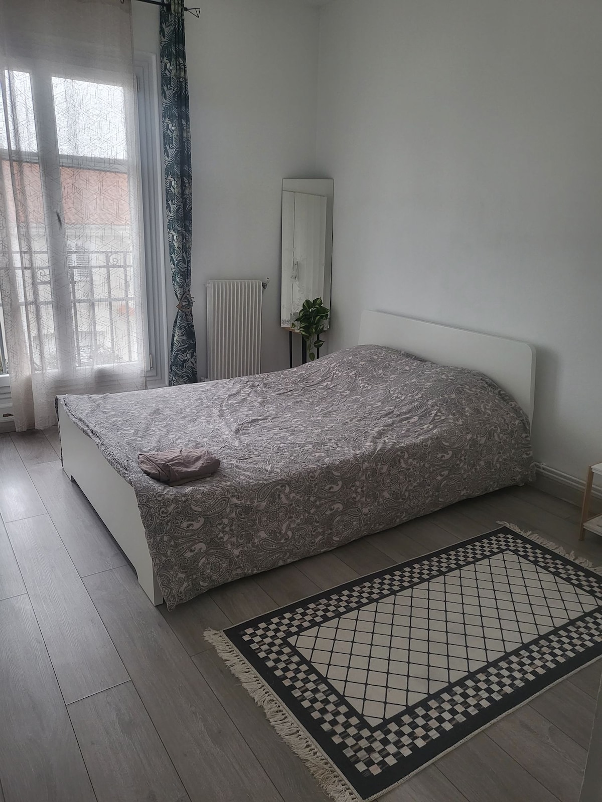 A bright sleeping area features a spacious bed draped in a subtle patterned cover. A mirror and a small plant are positioned beside the bed, while natural light filters through the sheer curtains of the window. A patterned rug adds texture to the wooden floor.