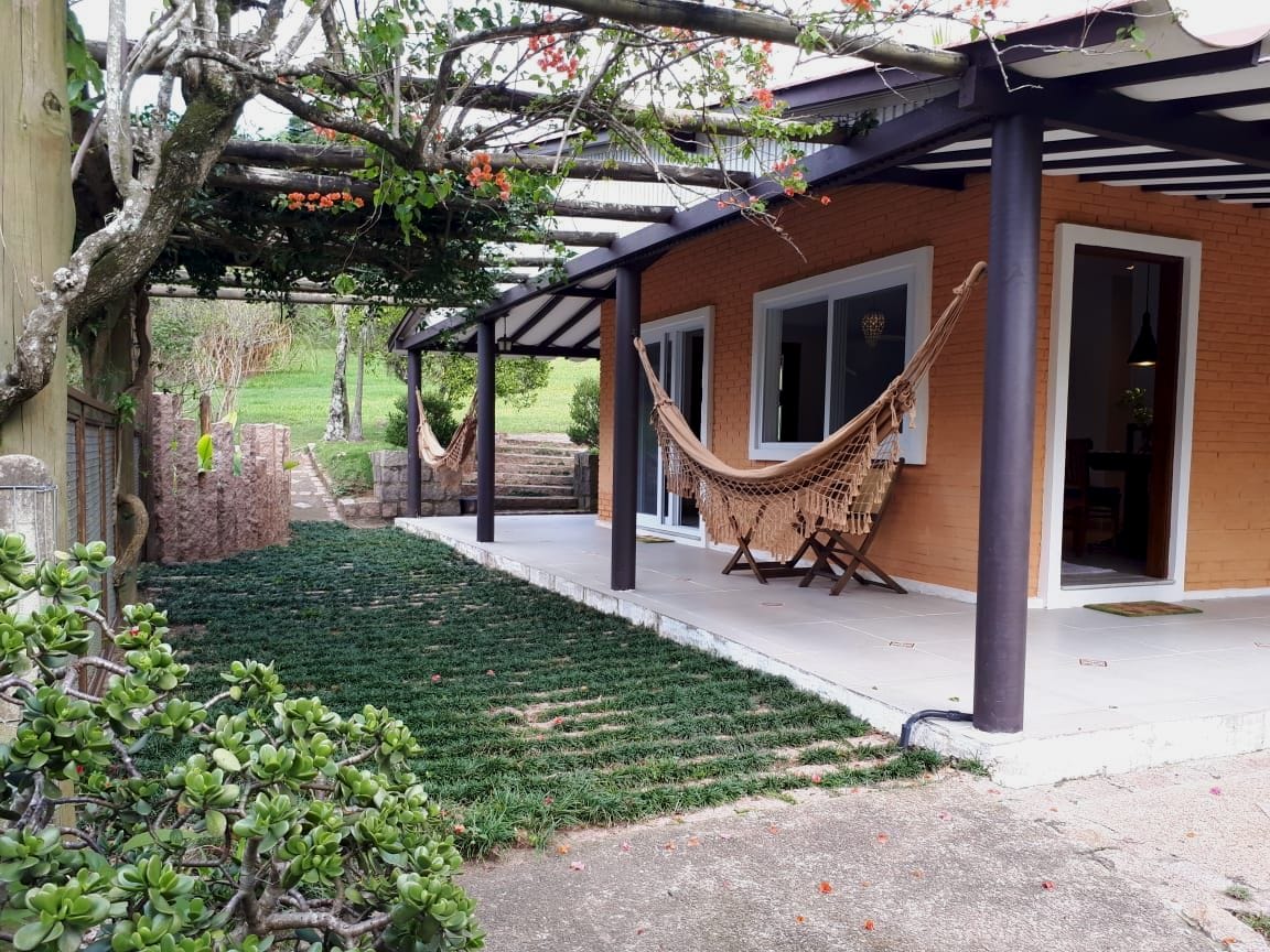 A covered patio area is featured with two hammocks hanging peacefully. A small table with folding chairs is positioned nearby. Lush greenery surrounds the space, complemented by flowering plants and a pathway leading to the well-maintained lawn.