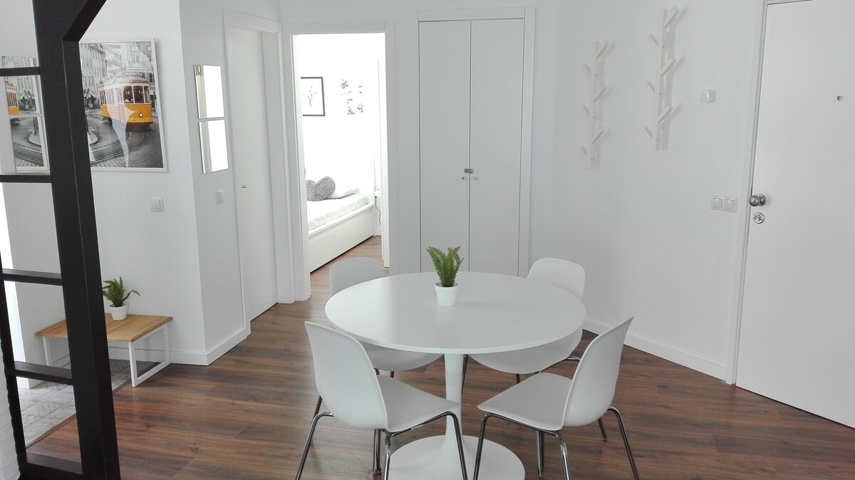 A light-filled dining area features a round table surrounded by four white chairs. A small potted plant sits at the center of the table. The space is connected to a hallway that leads to a bedroom, with a closet visible in the background.