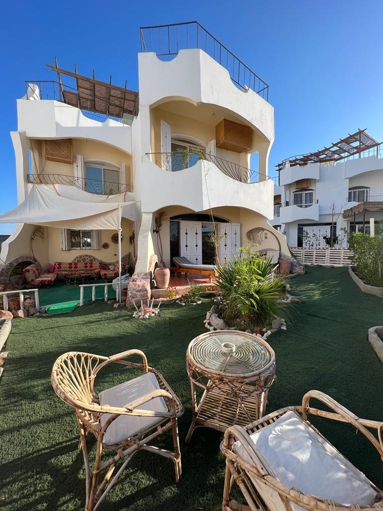 A charming outdoor seating area features woven chairs arranged around a glass table on a carpet of vibrant green grass. The villa's white facade and multiple levels are visible in the background, framed by clear blue skies.