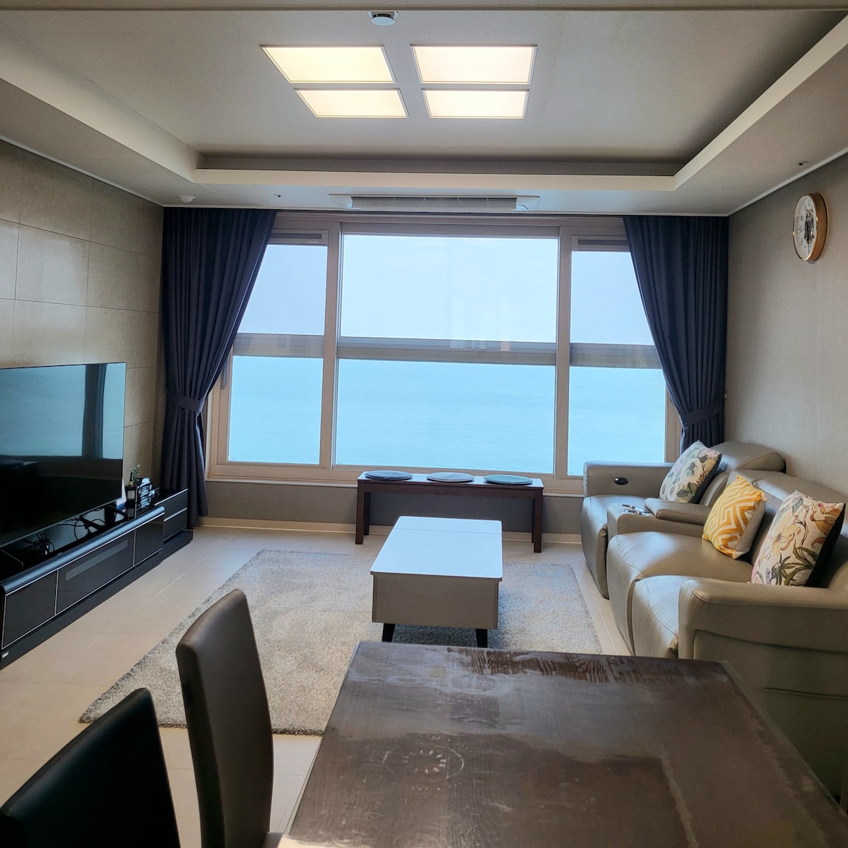 A spacious living area features a large window offering a clear view of the ocean. A comfortable sofa with decorative pillows is complemented by a simple coffee table. Soft lighting from the ceiling illuminates the room, enhancing the serene atmosphere.
