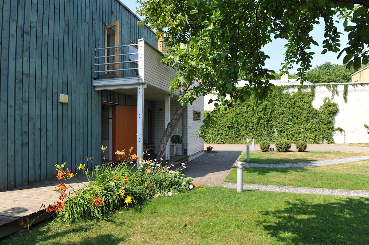 An outdoor view of the property features a well-maintained lawn with vibrant flower beds and a pathway leading to the entrance. The building showcases a wooden exterior, and greenery from nearby trees complements the serene environment.