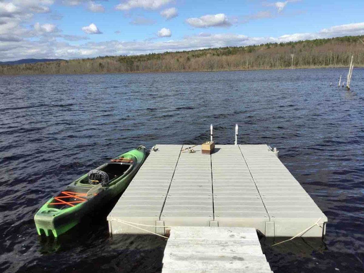 A spacious dock extends over the shimmering water, with a kayak resting alongside it. The calm surface reflects the sky, showcasing fluffy clouds and a backdrop of trees lining the shoreline.
