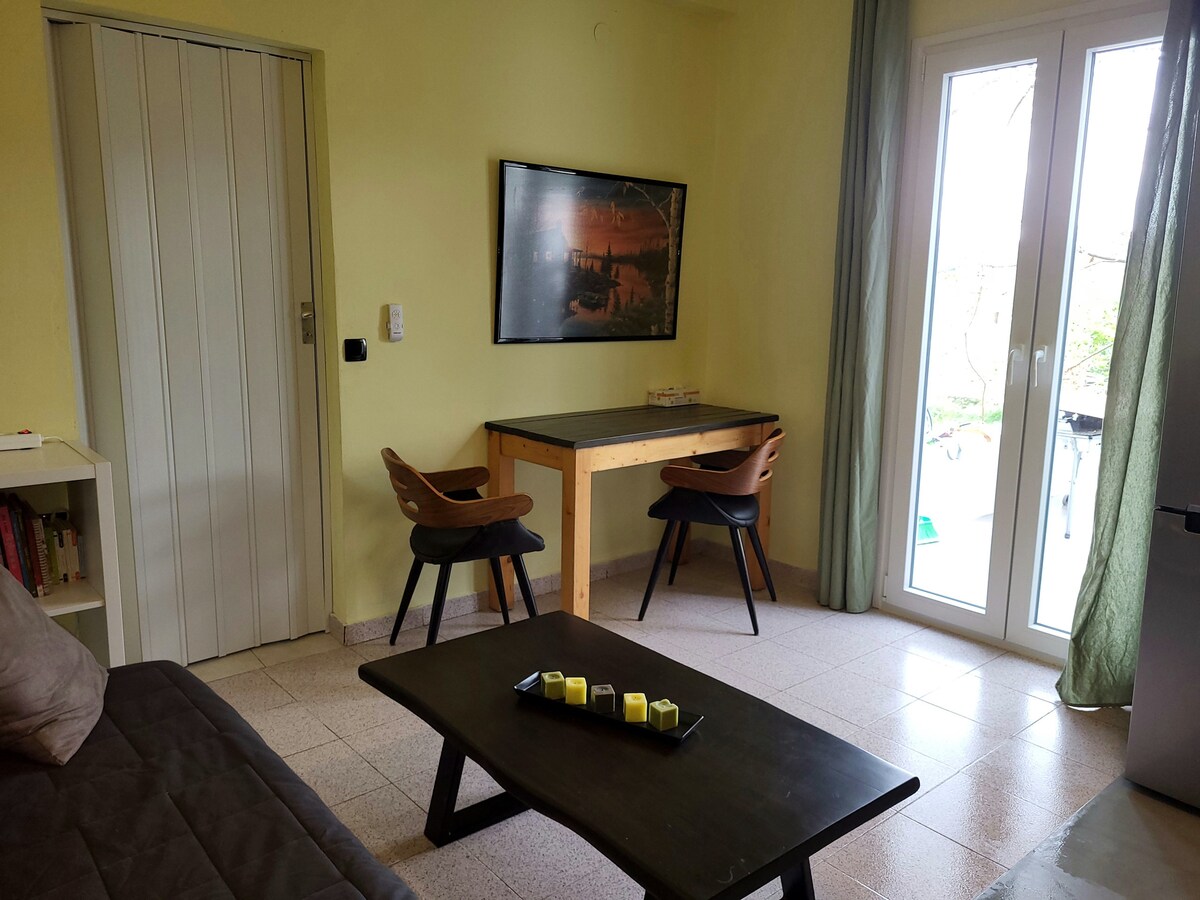 A bright seating area is visible, featuring a dark wooden coffee table complemented by a stylish sofa. Two chairs are positioned at a small desk against the wall. Large windows allow natural light to fill the space, with greenery slightly visible outside.