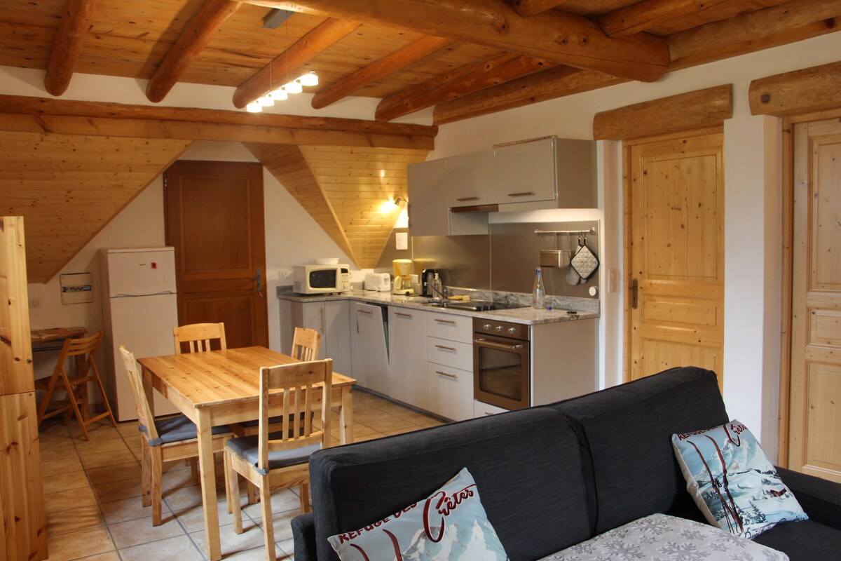 An open kitchen area is shown, featuring modern appliances, including a microwave and oven, set against wooden cabinets. A dining table with four wooden chairs is positioned nearby, complementing a comfortable seating area with a sofa adorned with decorative cushions. Exposed beams enhance the rustic charm.
