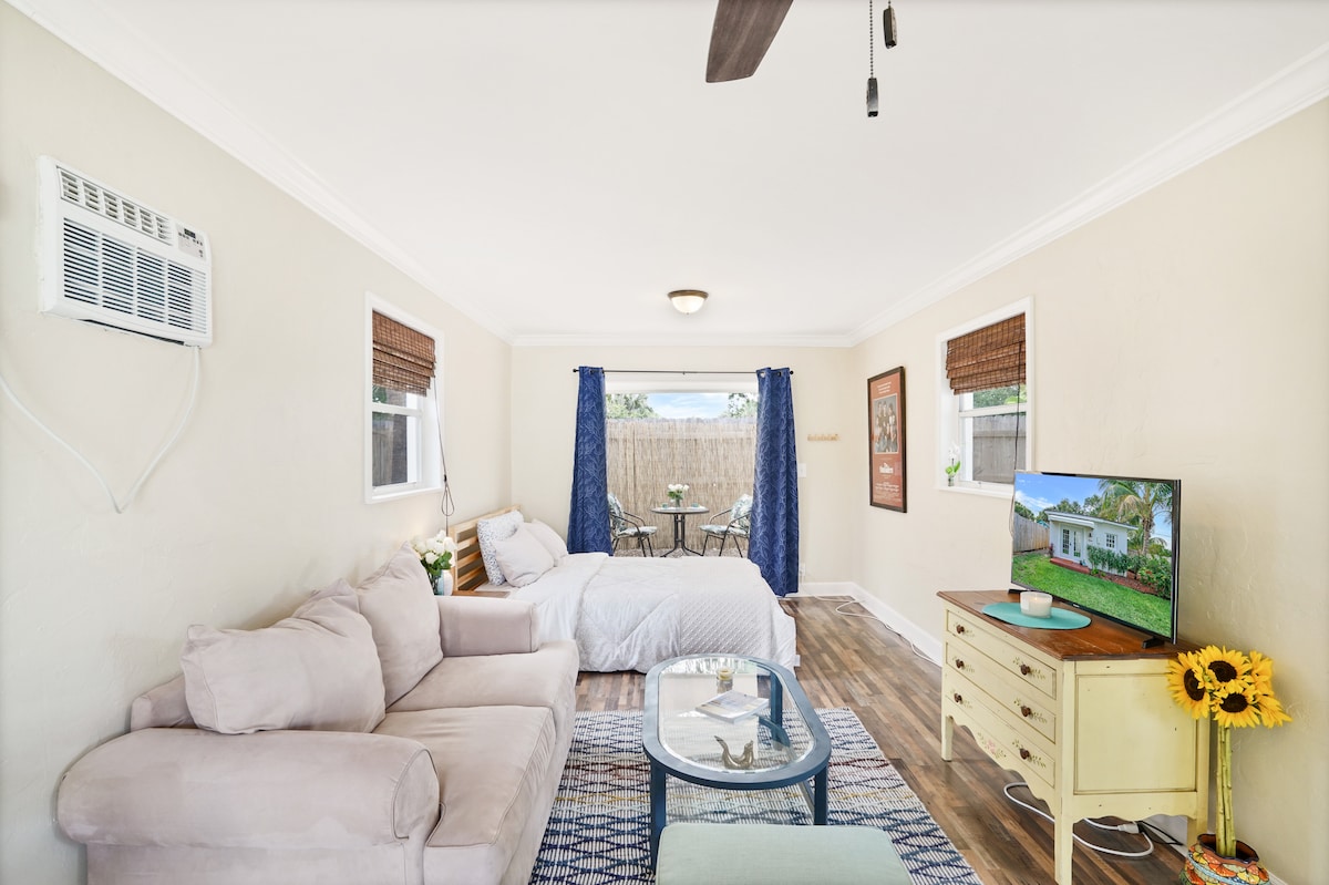 A cozy living area features a light-colored sectional sofa positioned beside a small coffee table. A spacious bed is visible in the background, accompanied by bright blue curtains that frame a window. An air conditioning unit is mounted on the wall, providing comfort.