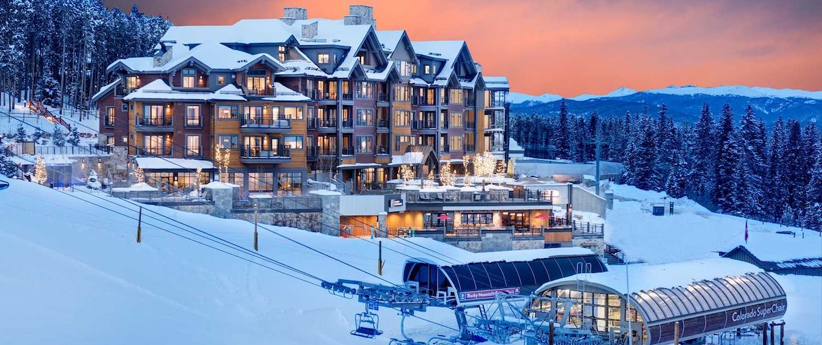 The exterior of the Grand Colorado on Peak 8 is depicted, featuring a large, multi-story building with a combination of dark wood and stone finishes. Ski lifts are visible, surrounded by snow-covered terrain and trees, with a vibrant sunset sky casting a warm glow over the scene.