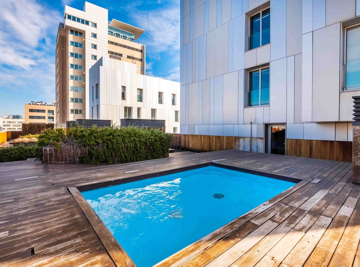 A private swimming pool is situated on a wooden deck surrounded by greenery. The pool's bright blue water contrasts with the light-colored building exteriors. Large windows provide a view of the modern urban landscape, contributing to a serene, inviting setting.
