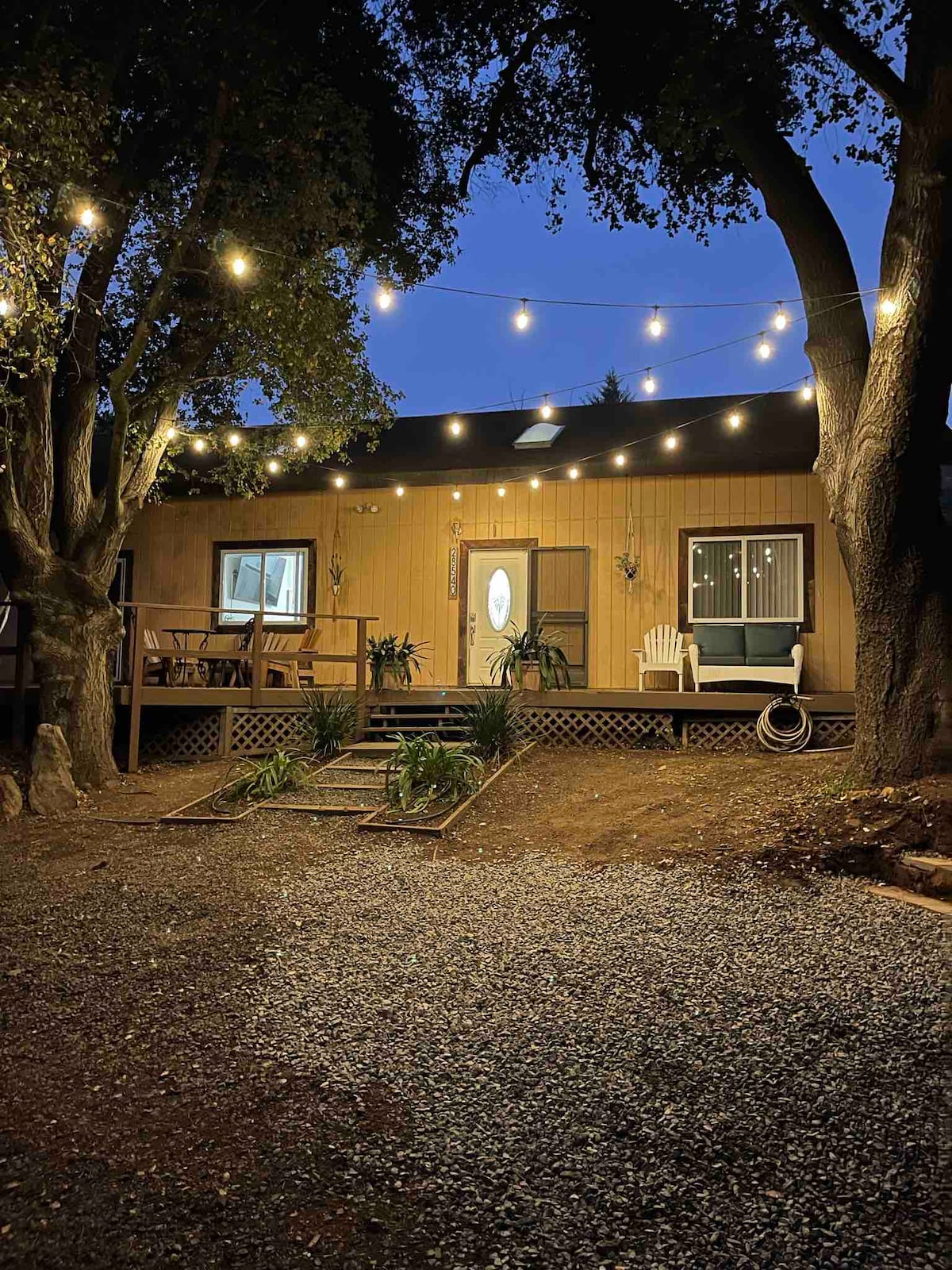 A rustic cabin is illuminated by soft string lights, creating a welcoming ambiance. The front porch features seating options and is surrounded by lush greenery. Gravel paths lead up to the entrance, framed by mature trees that enhance the tranquil setting.