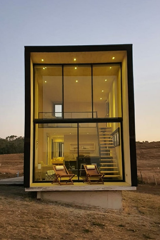 A modern two-story loft is presented, featuring large glass windows that illuminate the interior with warm light. Two wooden chairs are positioned outside, offering a view of the surrounding landscape. A staircase is visible on the left, leading to the upper level.