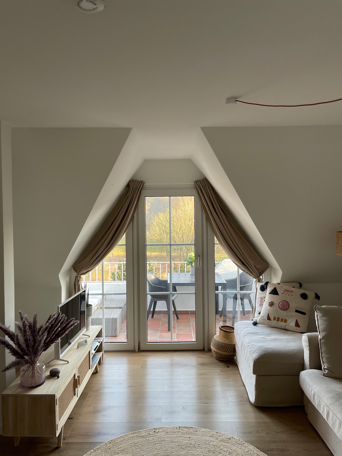A living area features a light-colored sofa situated beside a modern media console. The triangular window offers a view of the balcony, where outdoor seating is arranged. Soft curtains frame the window, enhancing the space's natural light and connection to the outdoors.