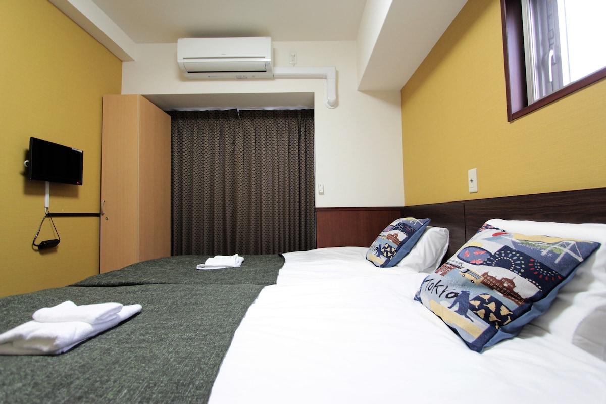 A well-organized bedroom features two spacious beds dressed in crisp white linens. Decorative pillows add a touch of color. A wall-mounted television is installed opposite the beds, while a window with dark curtains offers privacy. An air conditioning unit is visible on the ceiling.