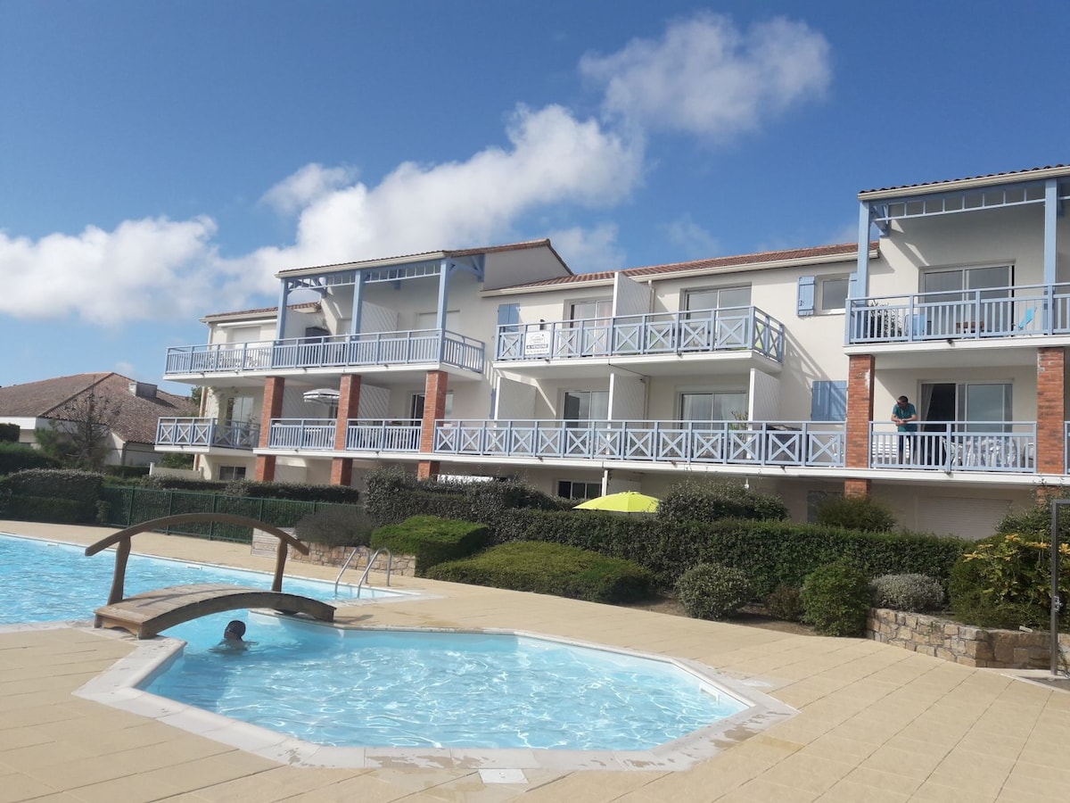 A three-story residential building features balconies with light blue railings facing a shared swimming pool. A small bridge leads to a circular area within the pool. Lush greenery surrounds the pool area, complemented by a partially cloudy sky.