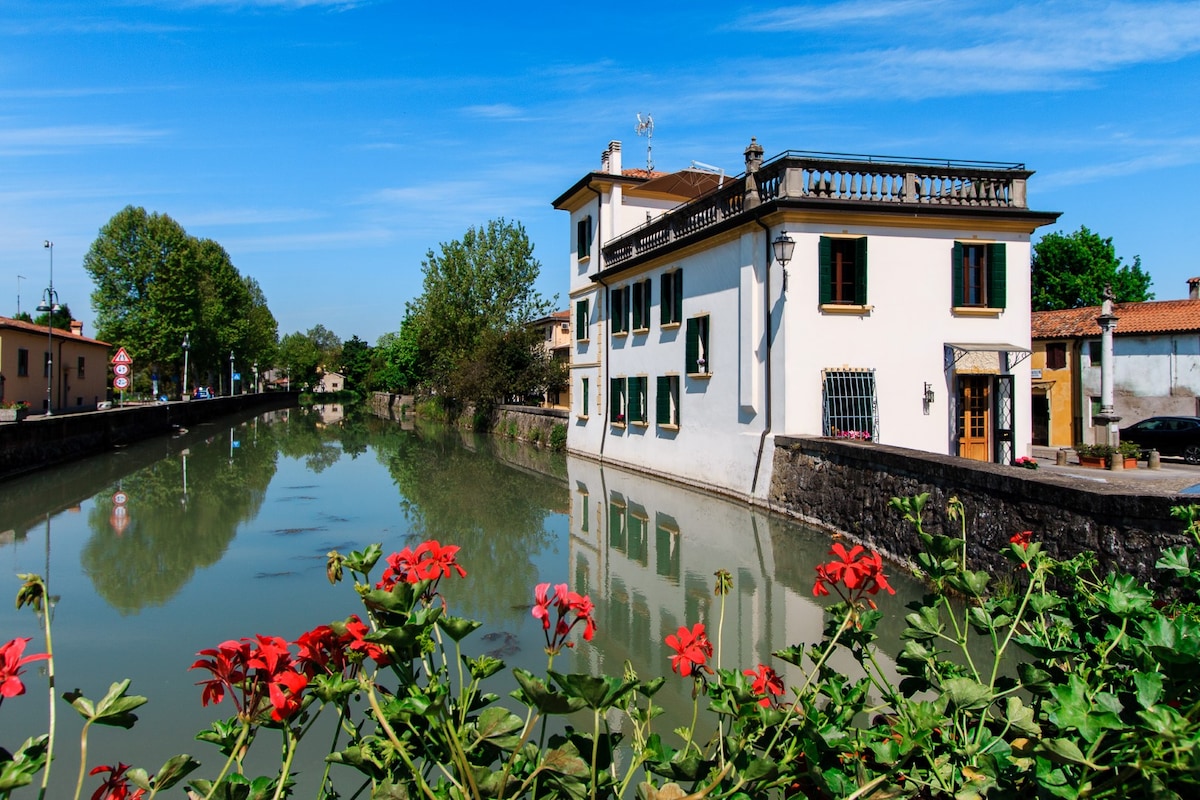 A charming building is reflected in the calm waters of a canal, flanked by vibrant red flowers in the foreground. Lush green trees provide a natural backdrop under a clear blue sky, enhancing the tranquil atmosphere of the location.