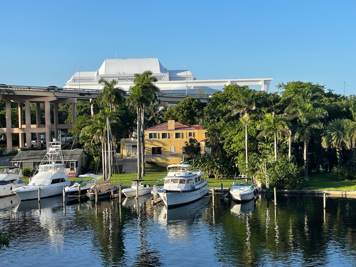 A tranquil scene along the Miami River features several boats docked along the wooden posts. Lush greenery and palm trees frame a historical yellow home with red roofing, while a modern structure is visible in the background against a clear blue sky.