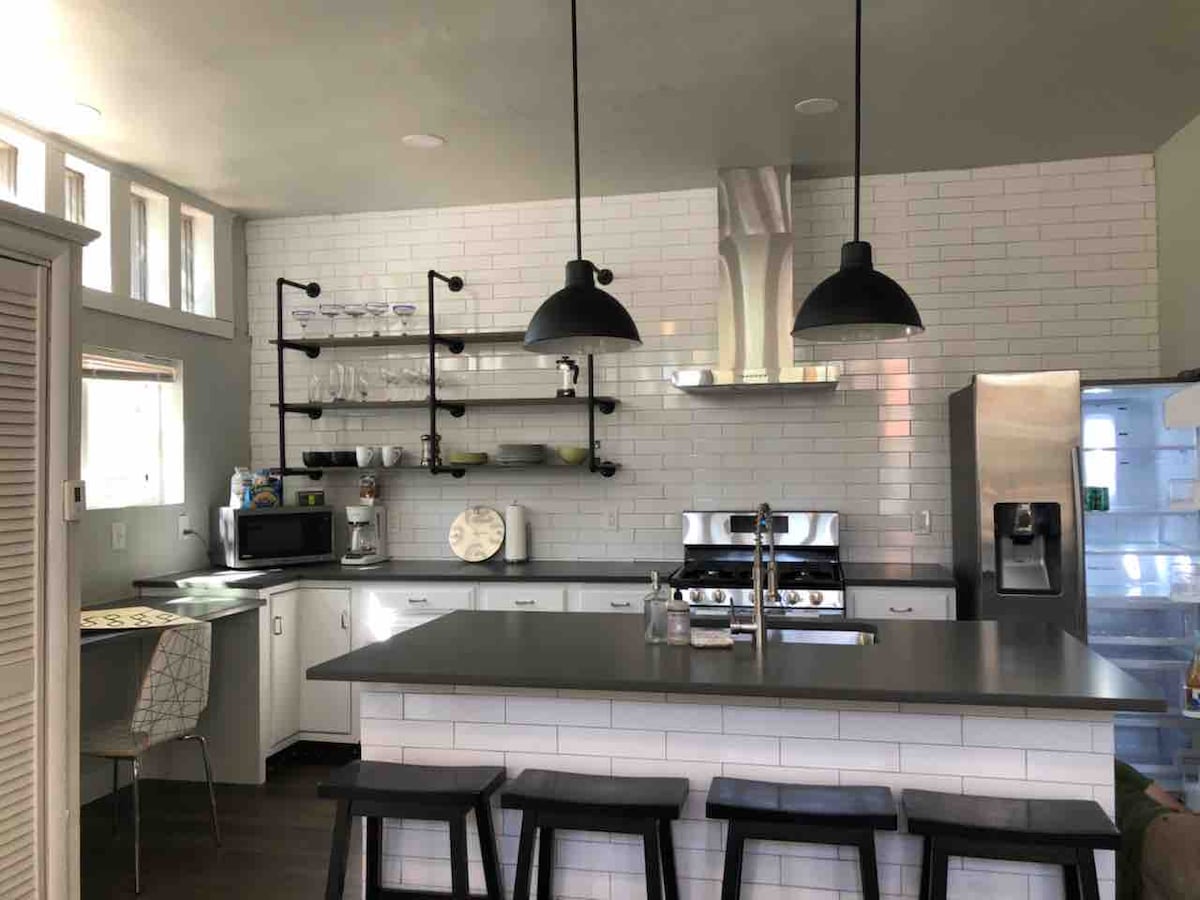 A modern kitchen is displayed, featuring stainless steel appliances and sleek granite countertops. Open shelving holds glassware and kitchen essentials. Industrial-style pendant lights hang above a spacious island with seating for three. Large windows allow natural light, enhancing the contemporary design.