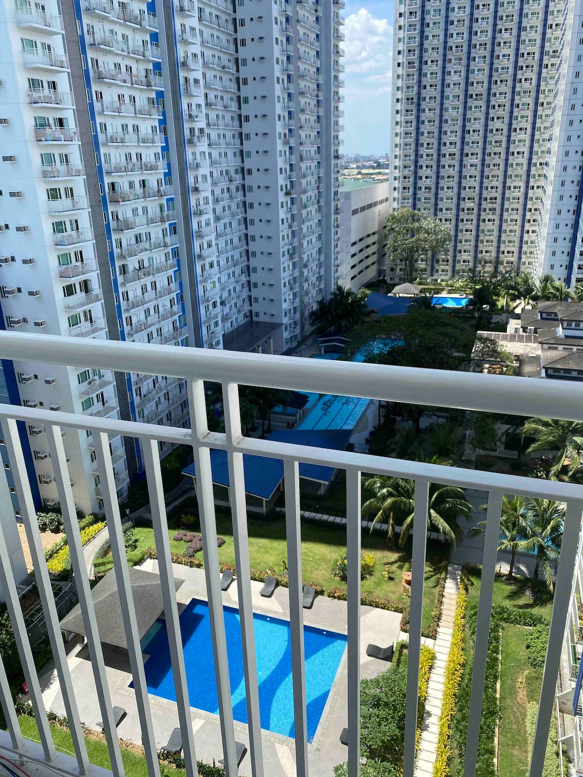 A view from the balcony presents a glimpse of community amenities, showcasing a blue swimming pool surrounded by landscaped gardens. Multiple high-rise buildings are visible in the background, with lush greenery interspersed throughout the property.