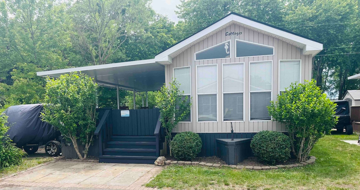 The exterior of a charming cottage is shown, featuring large windows and a covered porch. Lush greenery frames the entrance, which is accessible by a set of steps. A peaceful and inviting atmosphere is created by the surrounding trees.