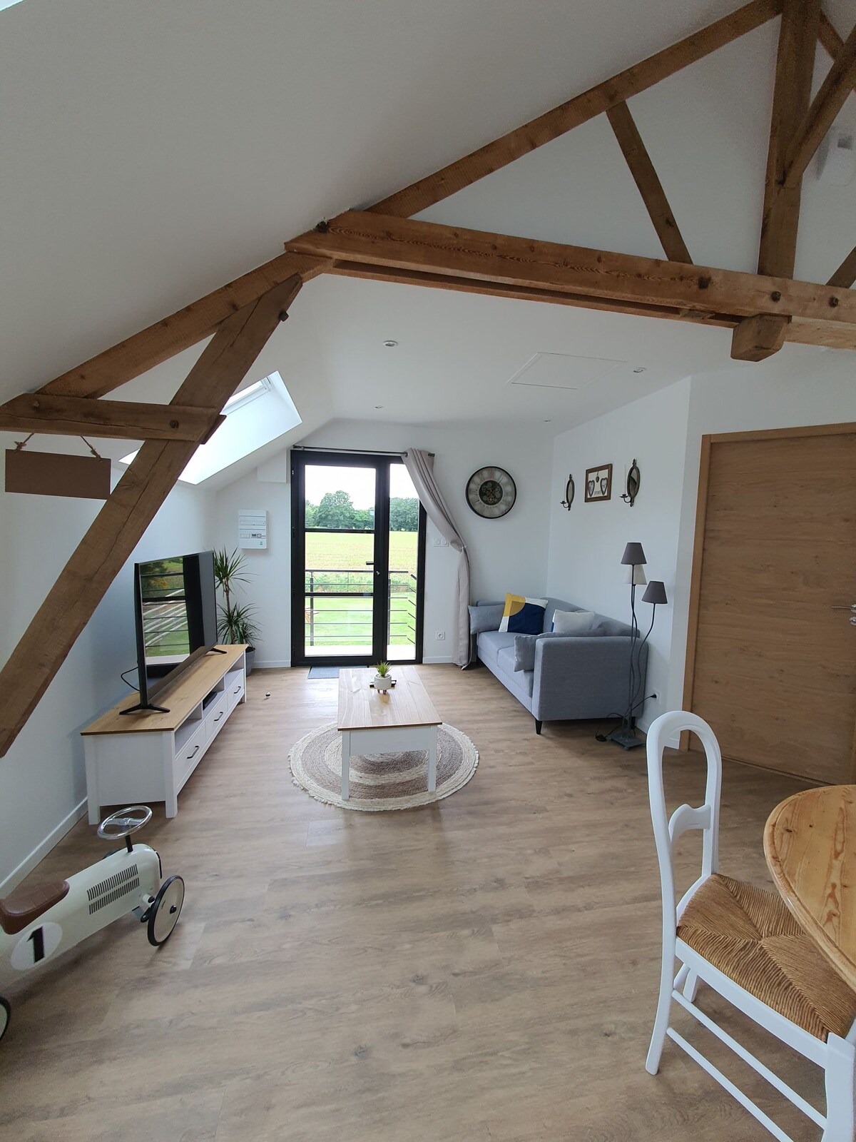 A light-filled living area features wooden beams, a comfortable gray sofa, and a television on a light wooden media console. The space is accented by a round area rug and offers views of the surrounding countryside through large glass doors. A decorative bicycle adds a charming touch.