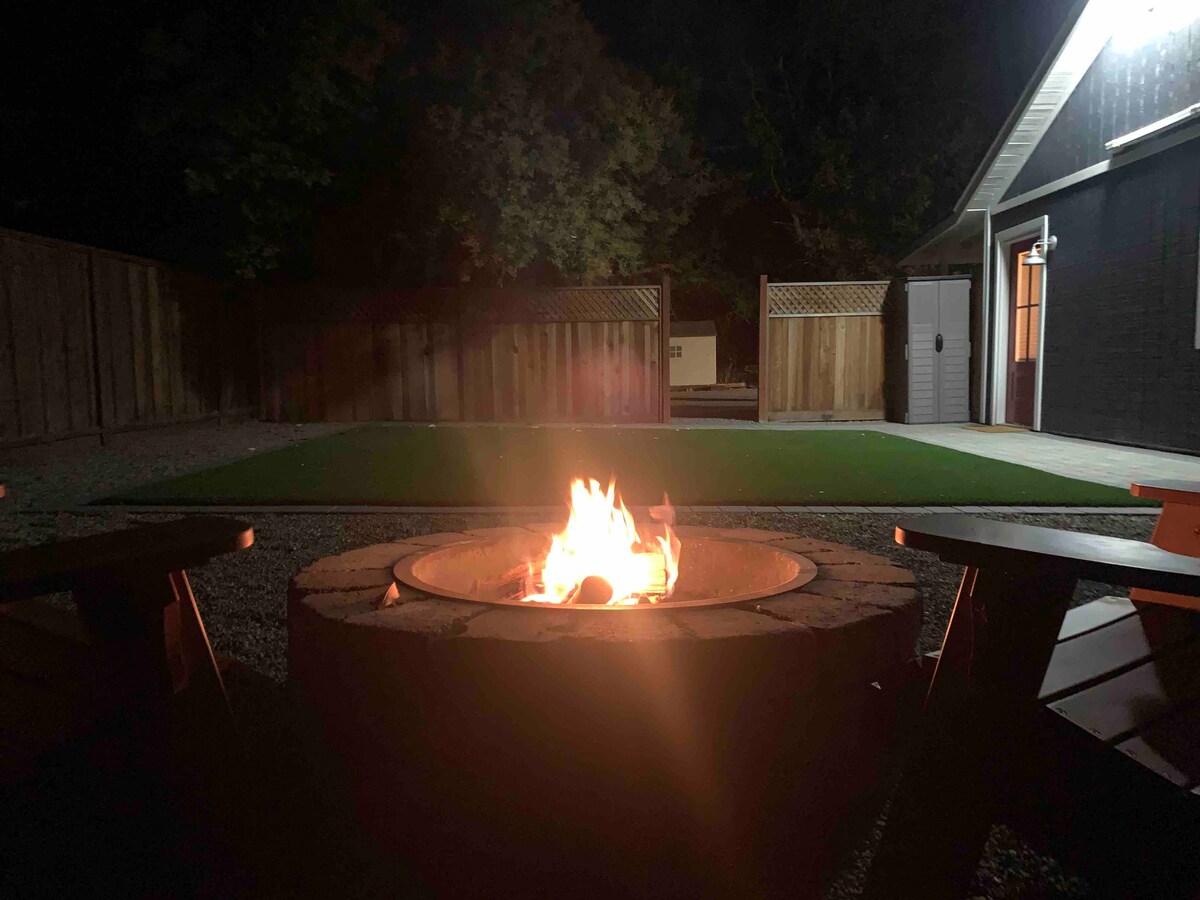 A stone fire pit emits a warm glow in the evening, surrounded by rustic wooden seating. The yard features a well-maintained grassy area and a wooden fence, providing a private outdoor space for relaxation or gatherings.