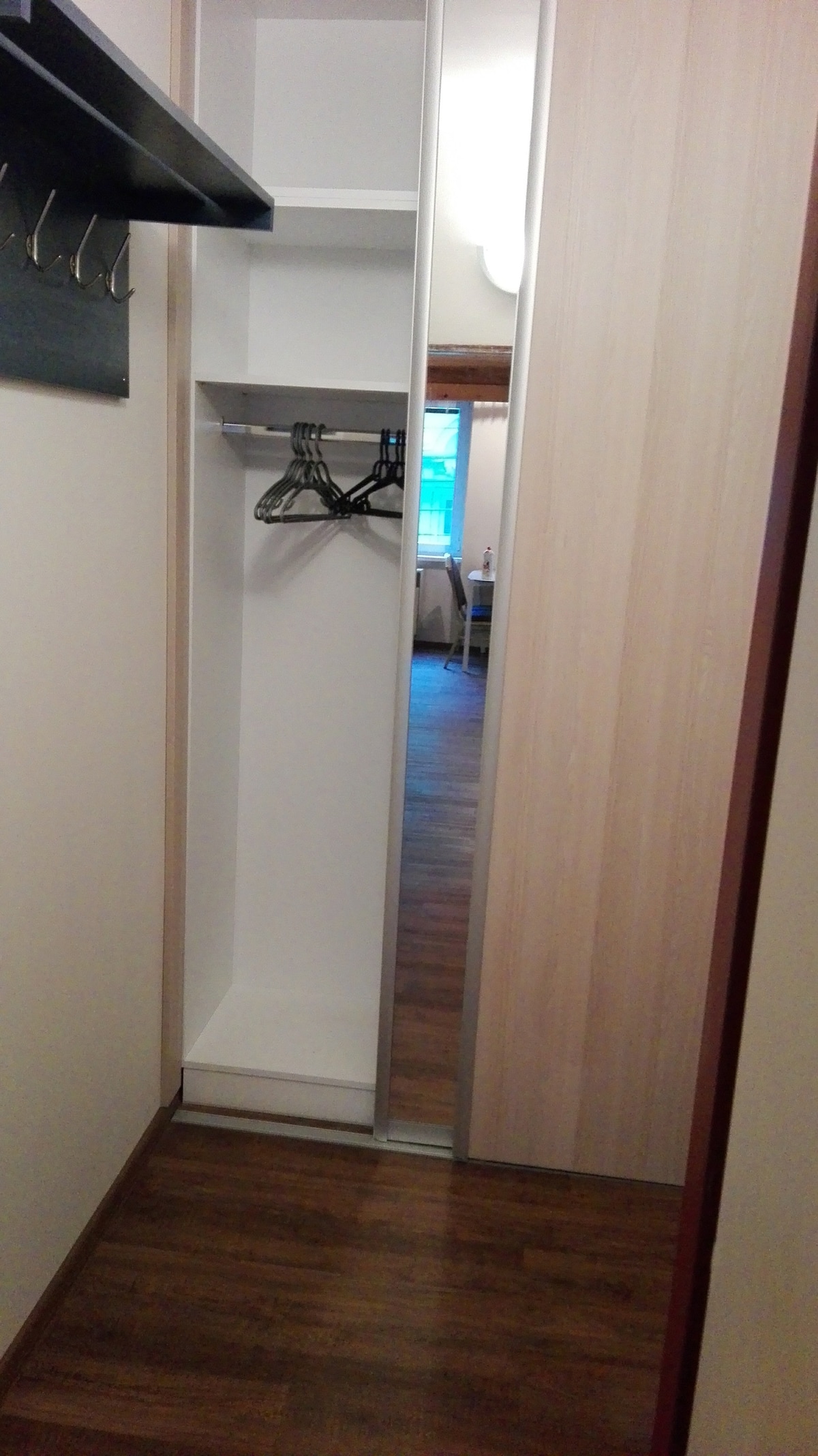 A compact closet area features light wooden panels and a full-length mirror. Two hangers are neatly arranged on a rod inside, with a small shelf above them. The space connects to a room with visible furnishings in the background.