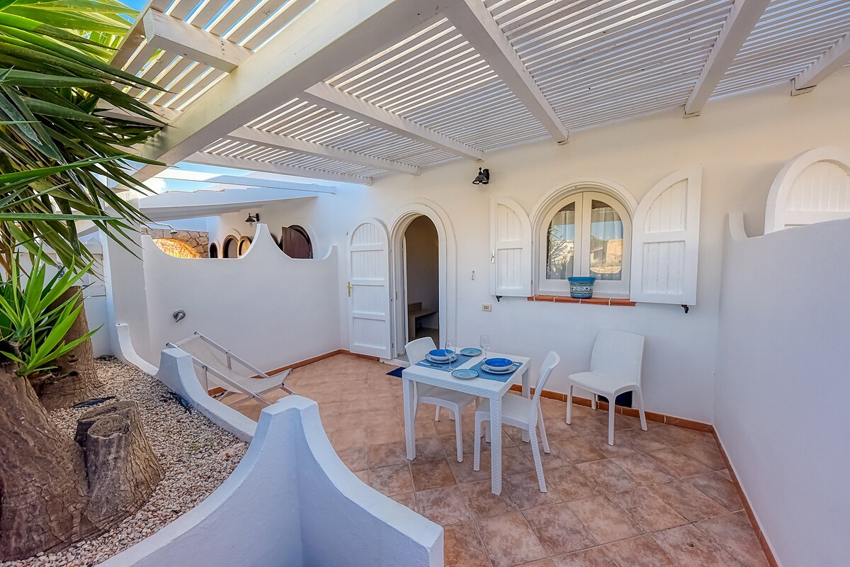 An outdoor veranda features a white table set for dining, accompanied by four chairs. A sun lounger is positioned nearby, with a tree and decorative stones adding a touch of greenery. Arched windows and double doors provide access to the interior.