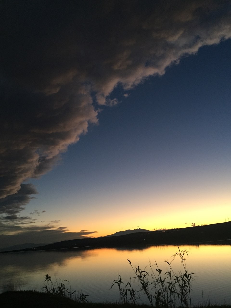 A serene lake landscape features a calm water surface reflecting the gradient colors of twilight, shifting from deep blue to warm orange. Dark clouds hover above, contrasting with the vibrant horizon line. Silhouettes of reeds are visible at the water's edge.